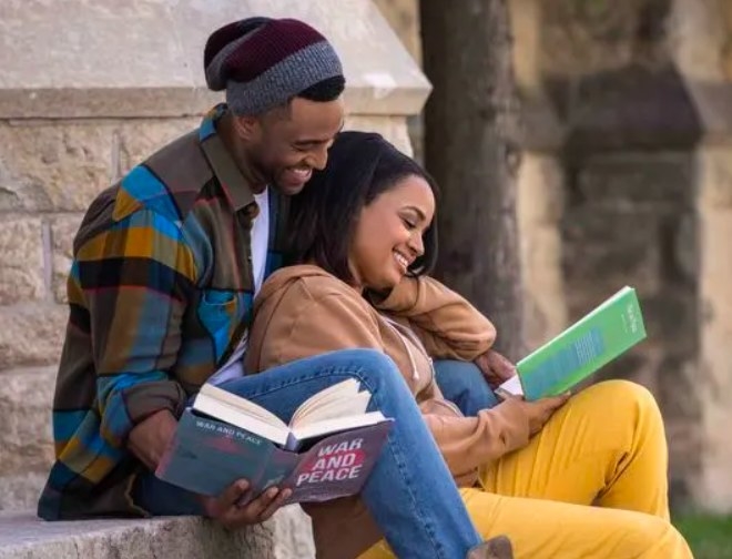 Kyla Pratt, Brooks Darnell sit cuddled together on an outdoor stone structure, reading books and smiling