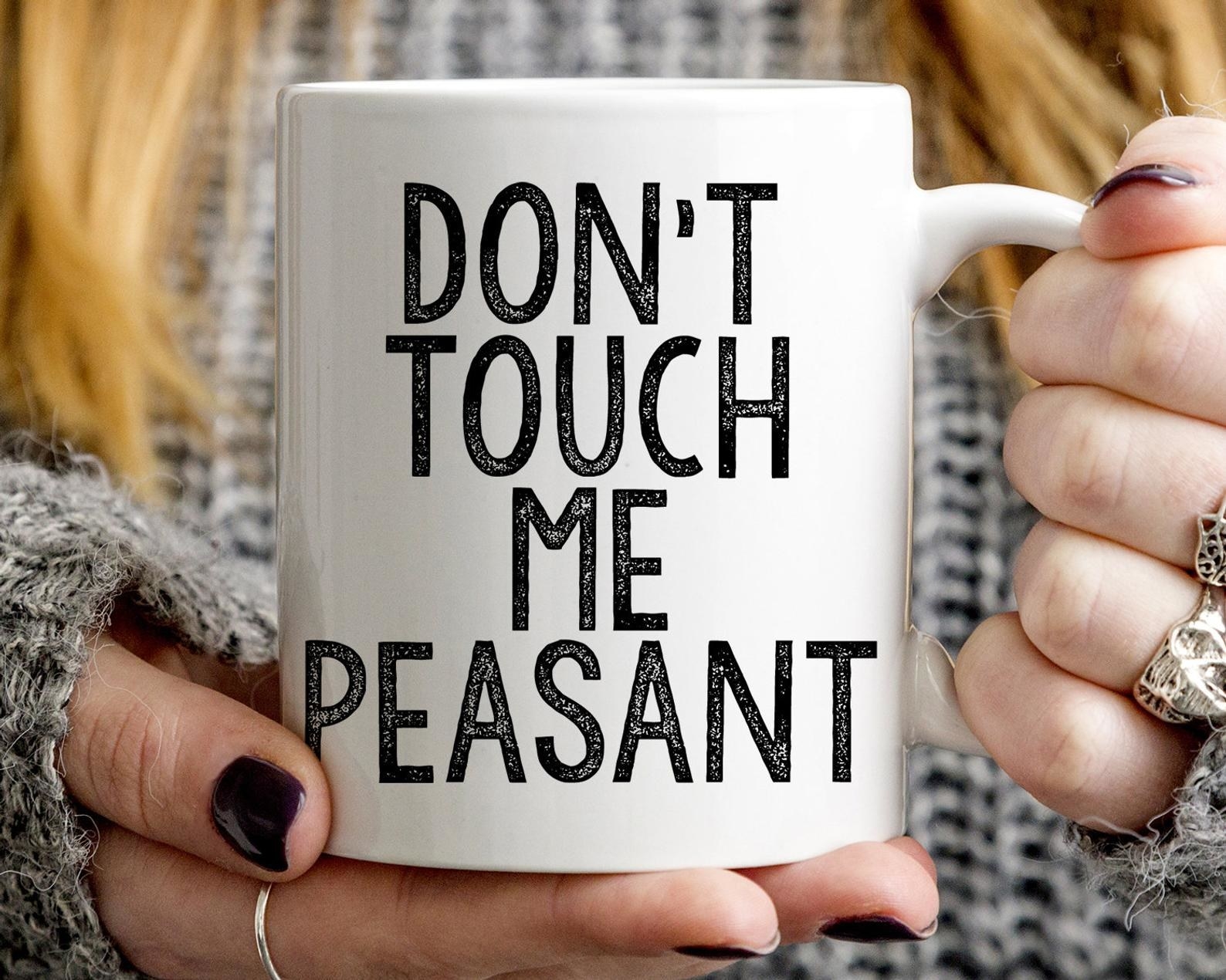 a person holding the white mug that reads &quot;don&#x27;t touch me peasant&quot; in black capital letters