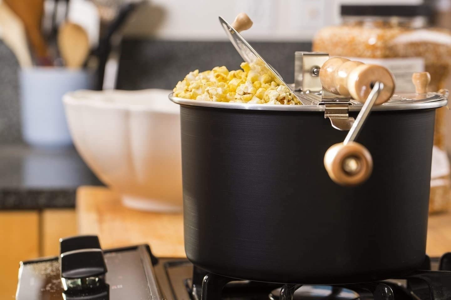 The popper, which is a black pot with a silver lid and wooden hand crank, on a stove