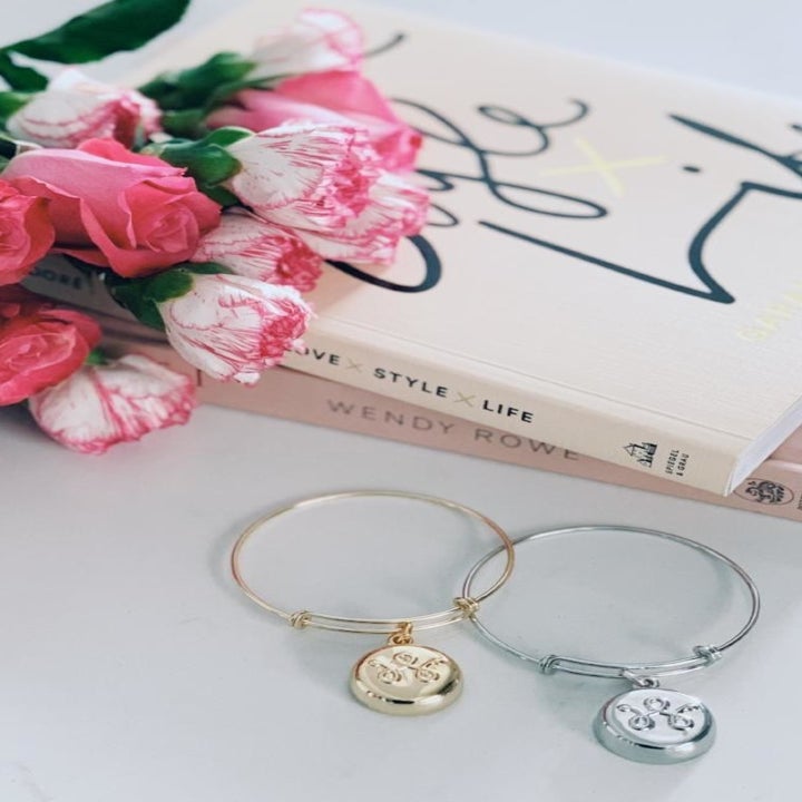 A silver and gold invisaWear bangle with a round charm sitting next to a stack of books and a bouquet of flowers 