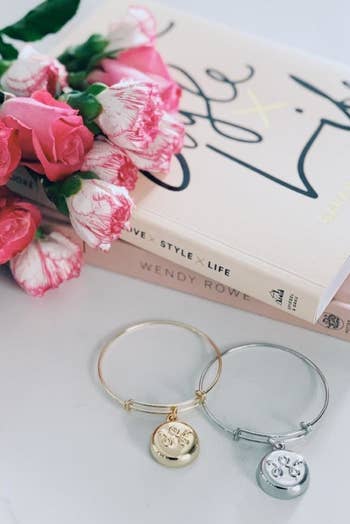 A silver and gold invisaWear bangle with a round charm sitting next to a stack of books and a bouquet of flowers 