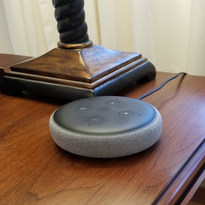 A reviewer photo of a charcoal Echo Dot sitting on a side table next to a lamp 