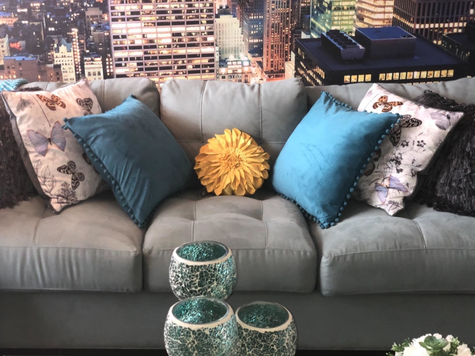 Sunflower accent pillow wedged in between blue pillows and butterfly-pattern pillows on a light gray couch