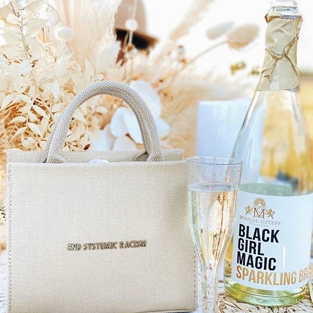A bottle of black girl magic sparkling brut photographed outdoors next to a champagne flute and a small beige bag that read end systemic racism