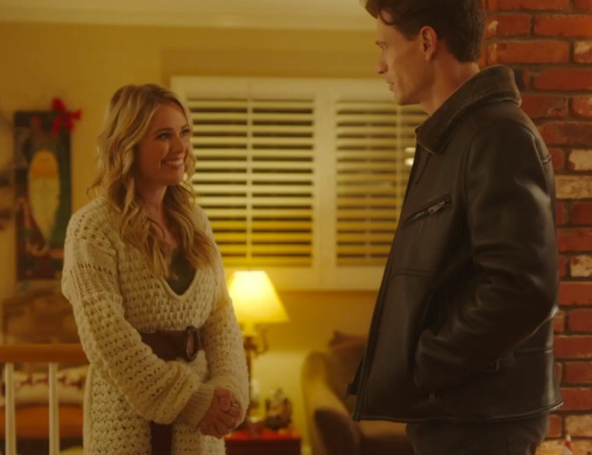 Natalie Hall and Jon Prescott stand in a living room facing each other and smiling