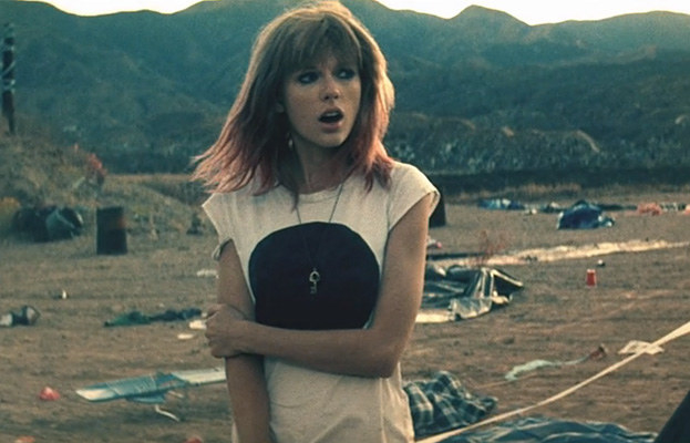 a woman with bangs and shoulder-length hair stands in a field in front of mountains. she has her mouth open, mid-song, looking to the left. she wears a t shirt and long necklace, and left arm is bent in front of her holding her right arm