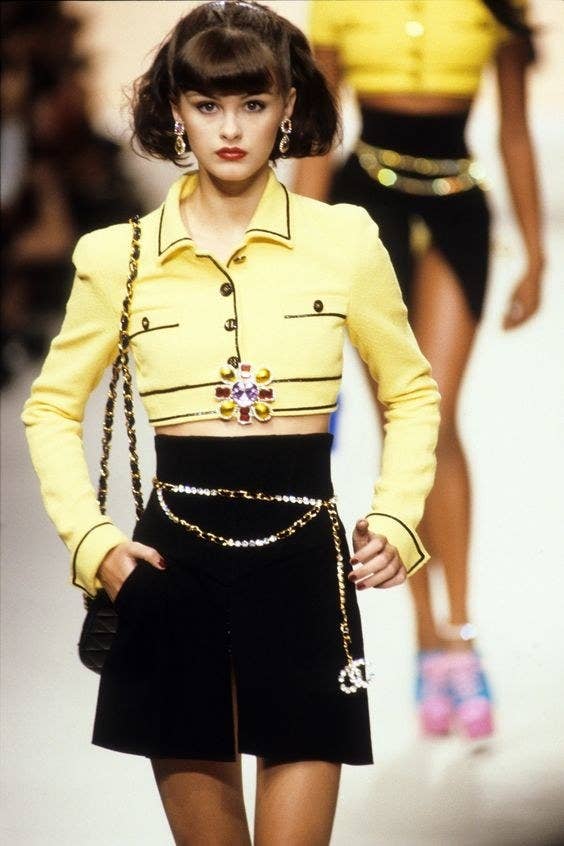 Chanel Spring 1995 Ready-To-Wear; Yellow blazer with black detail, black skirt, black bag, gold and silver Chanel chain, red, purple and yellow flower pin.