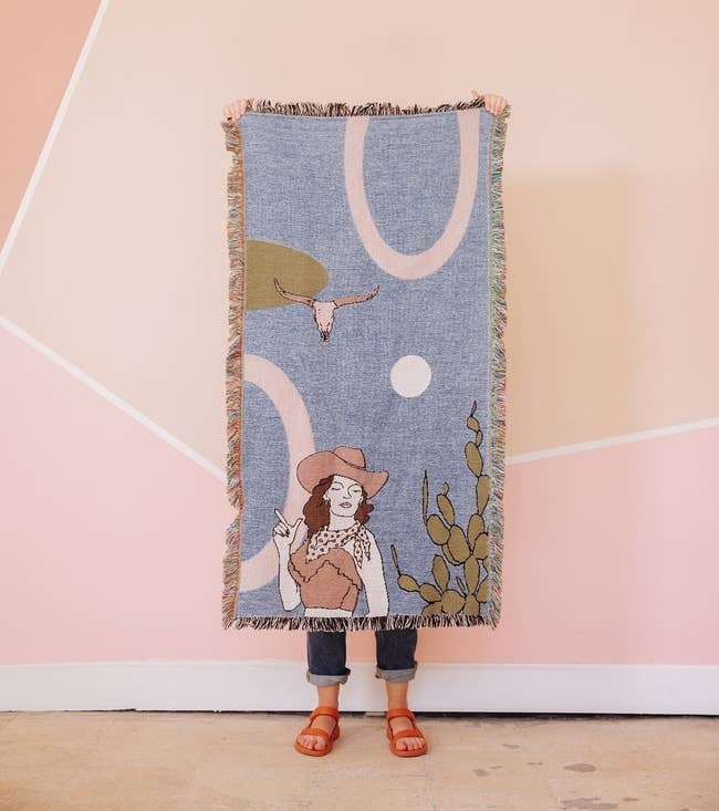 A rectangular throw blanket with a cowgirl, cactus, and animal skull on a minimalist geometric background