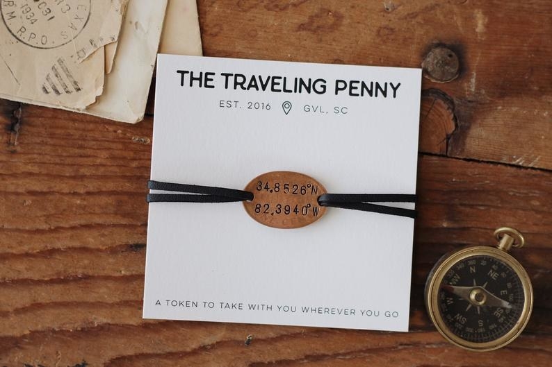 A bracelet with a dual leather brand and a copper charm engraved with coordinate wrapped around packaging that reads &quot;The Traveling Penny, EST 2016, GVL, SC&quot; 
