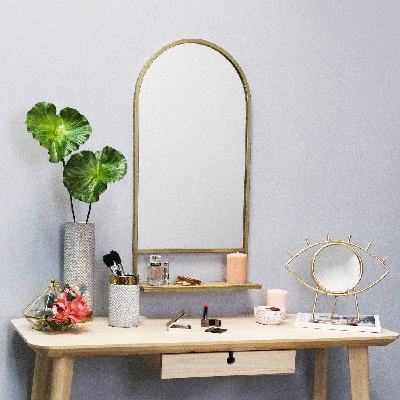 The mirror, which has a gold-toned rim, an arched top and rectangular bottom, and a collapsible shelf on the bottom