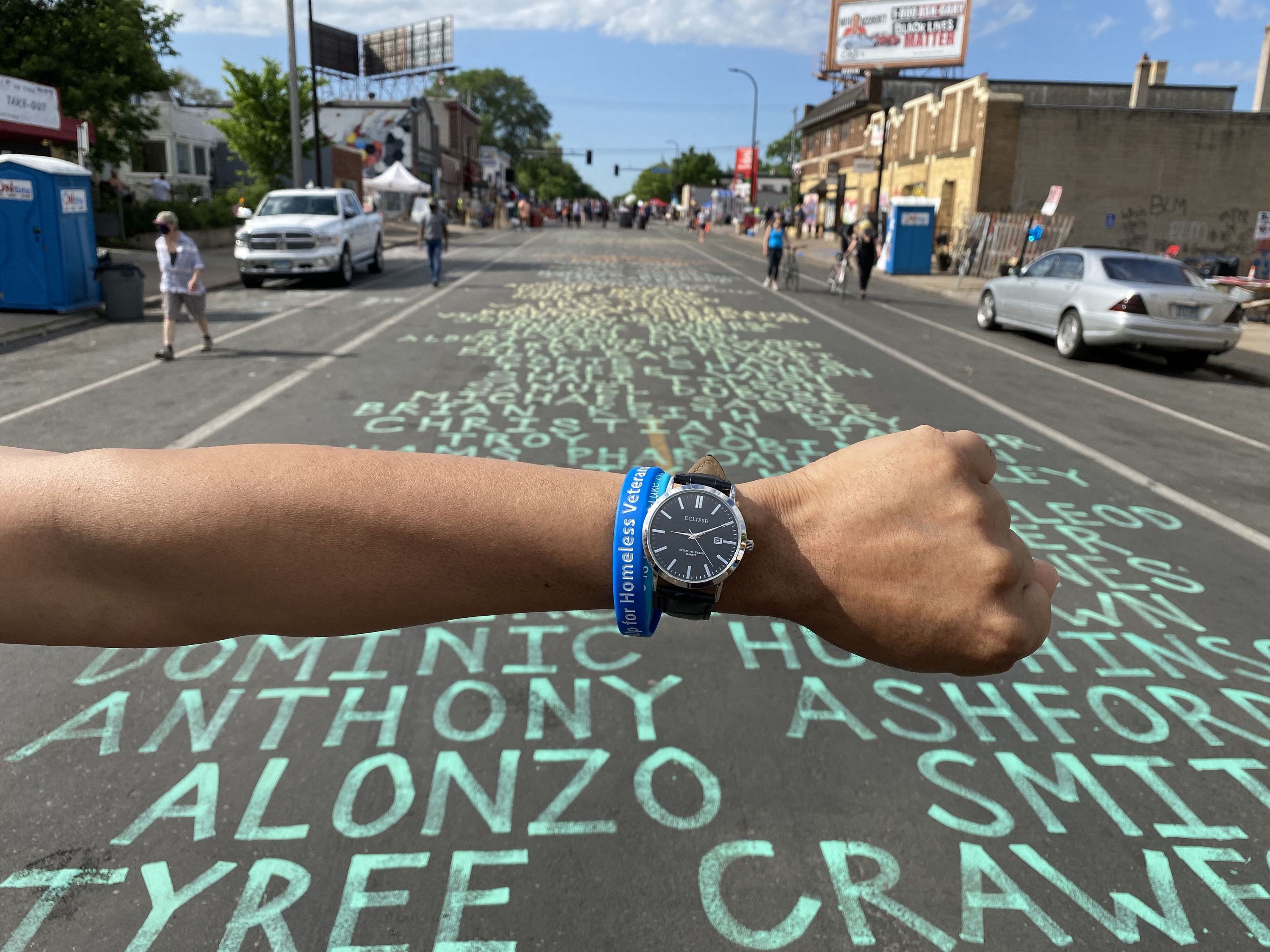 An arm wearing a watch in front of a list of names written in chalk on the street