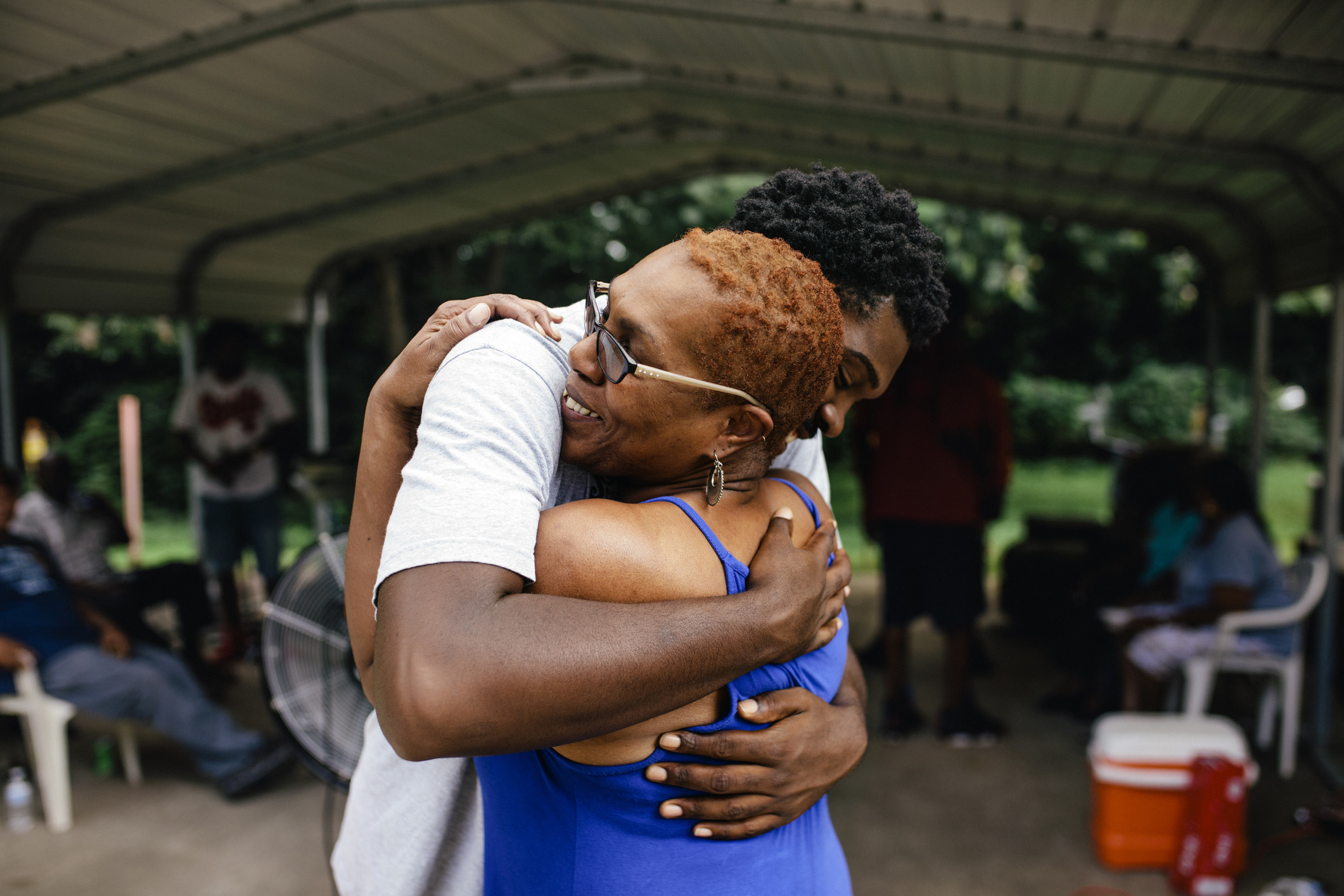 Middle aged black mother hugging her young adult son. Family outdoor cookout happening in the background. 