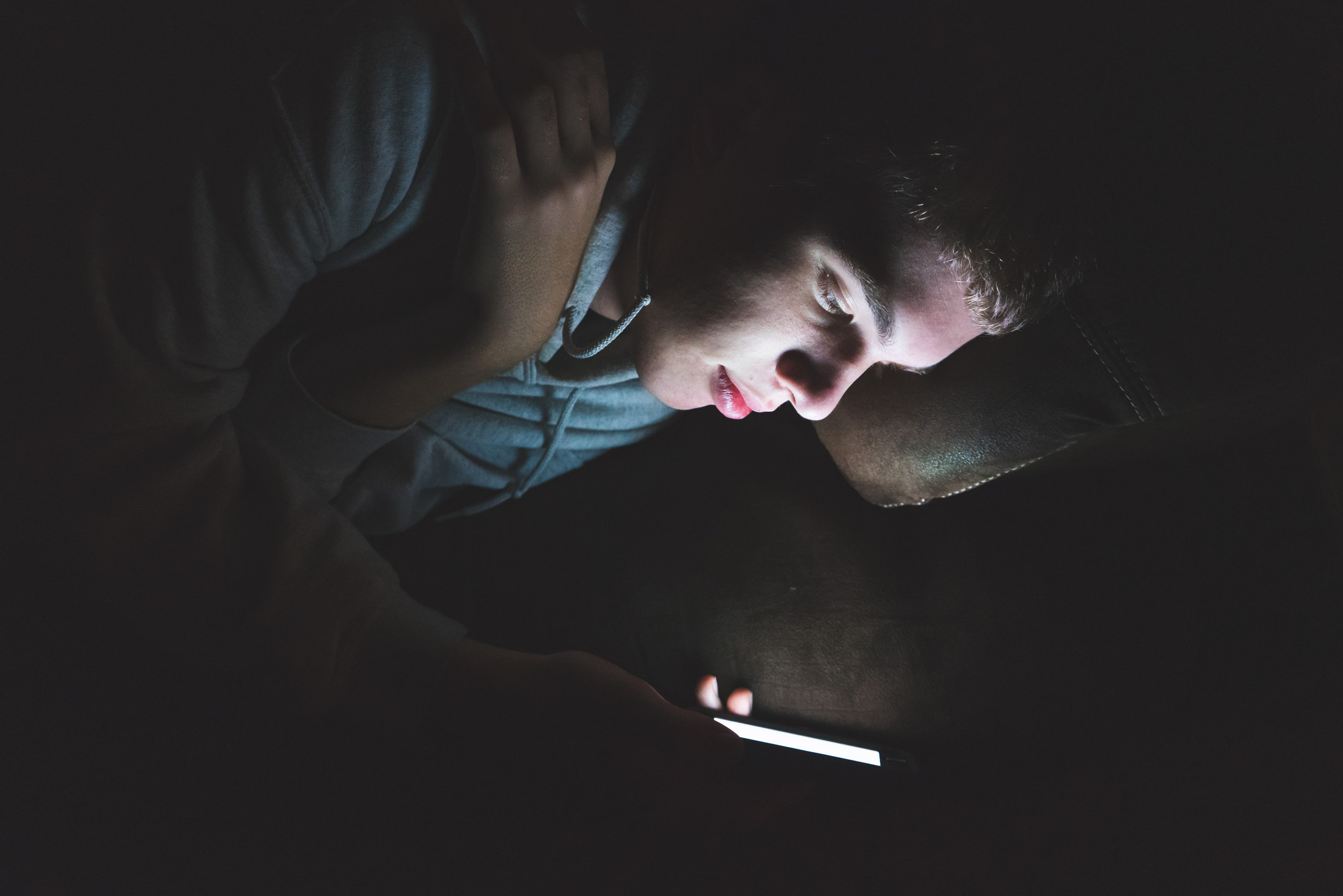 The image displays a teenager lying down on a couch in the dark. The light from the screen of his smartphone is illuminating his face.