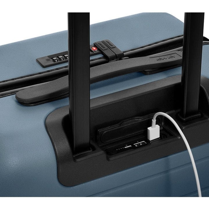 A closeup of the top of the carry-on suitcase showing a top handle and locking mechanism and a charging cor plugged in 