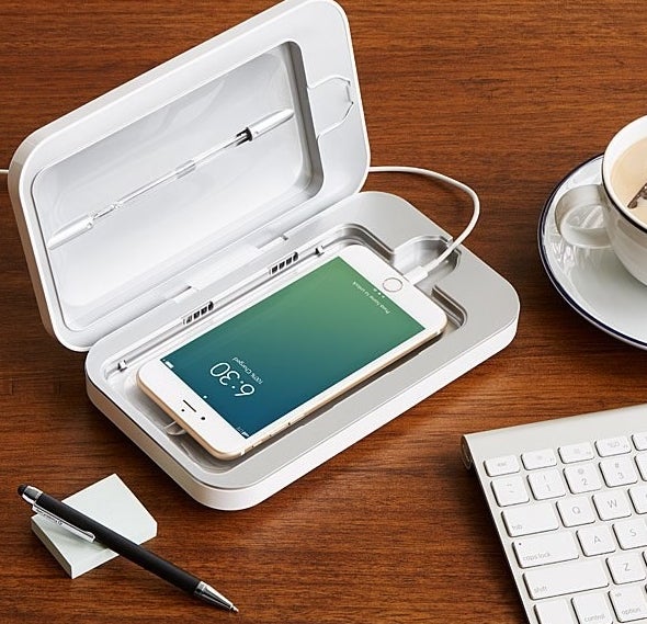 A smartphone sitting in the open UV phone sanitizier, sitting on a desk next to a keyboard and pen