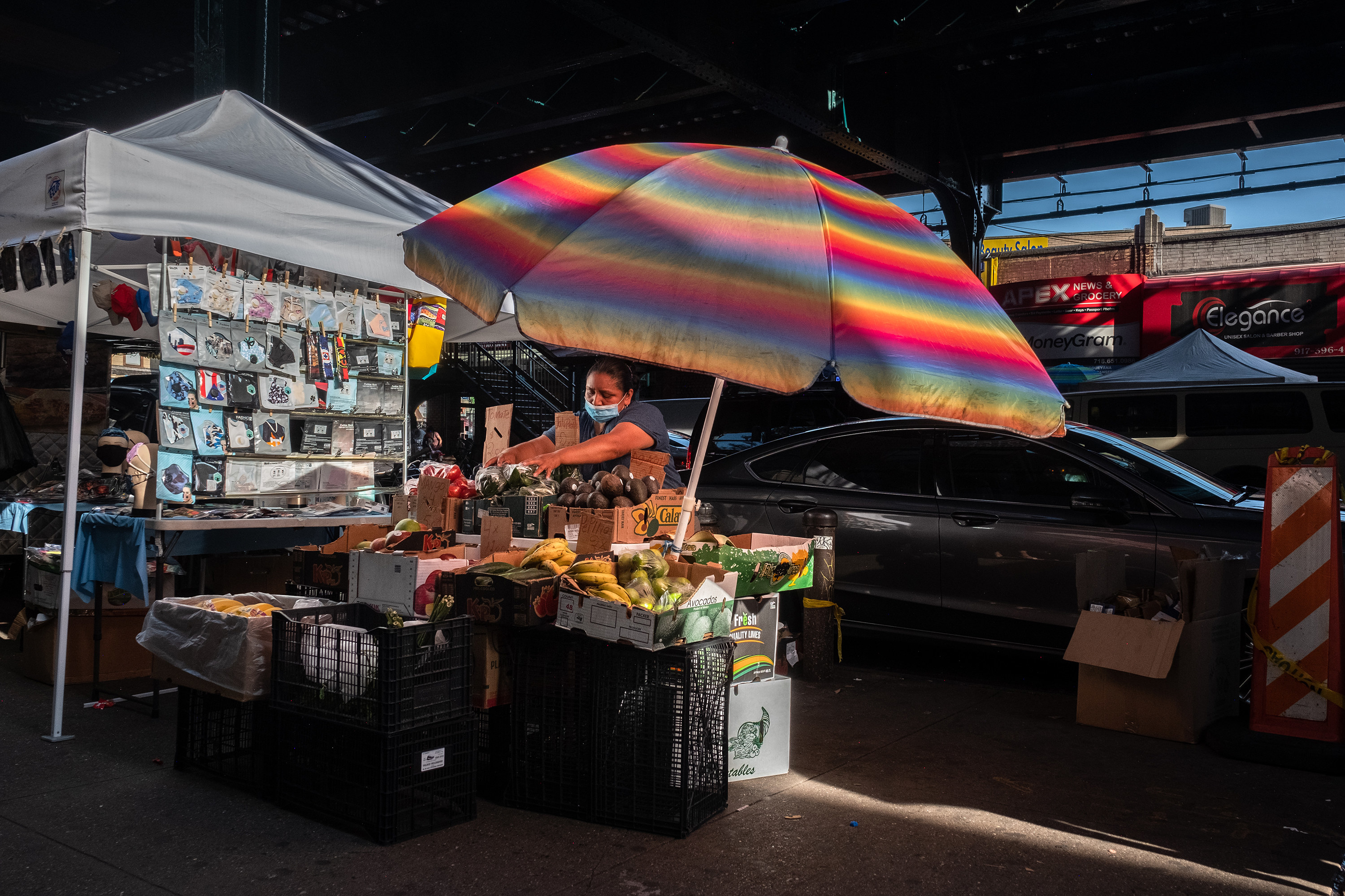 A woman arranging vegetables underneath a brightly colored umbrella underneath the an elevated subway platform in New York. 