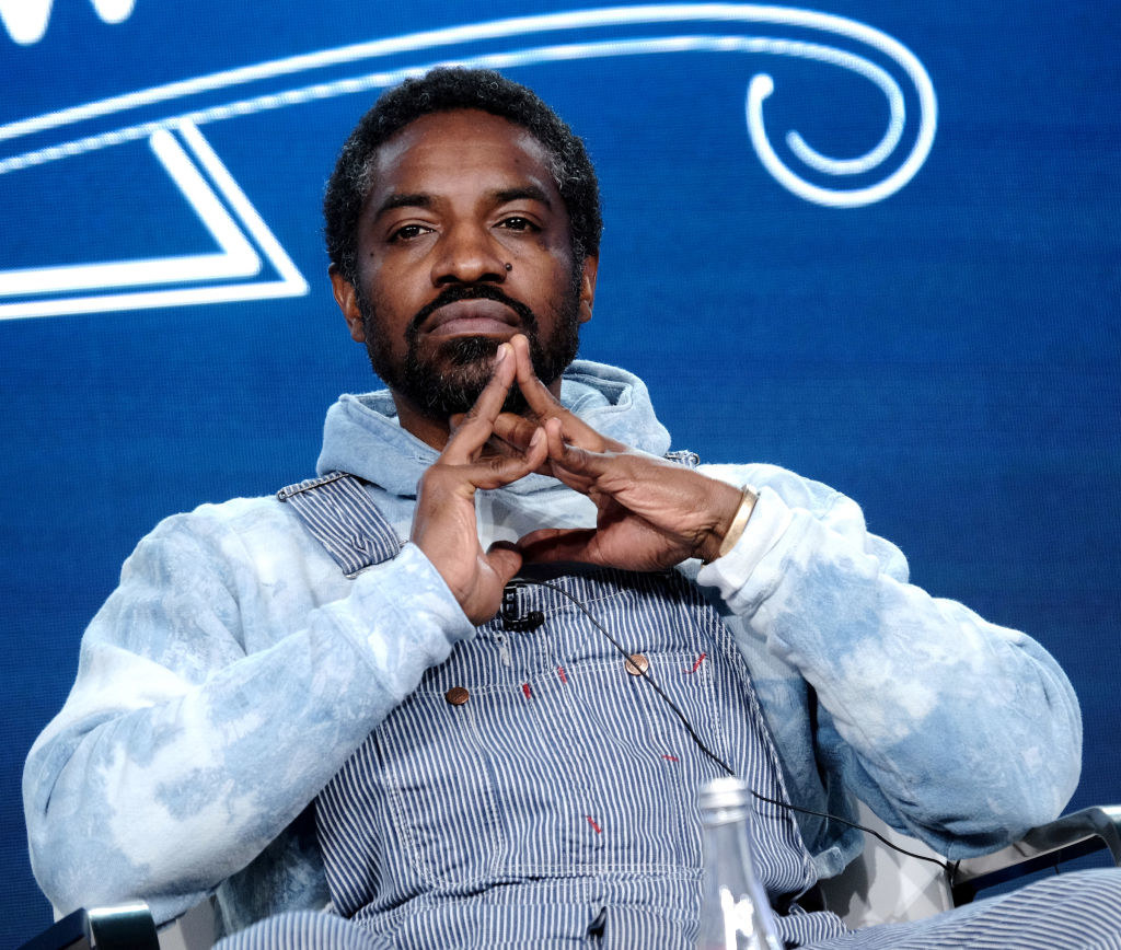 André 3000 sitting in a chair while wearing overalls, clasping his hands together while thinking pensively