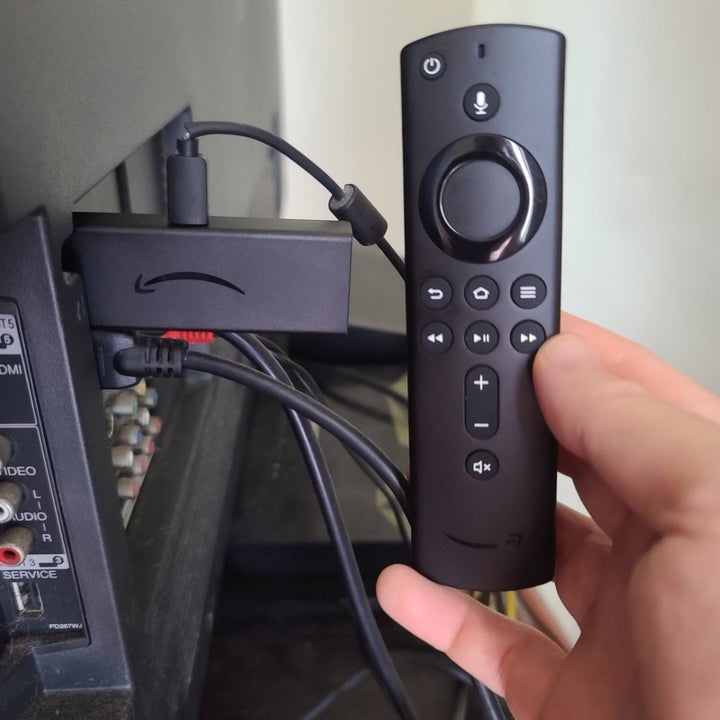 A reviewer photo of the Fire Stick plugged into a TV and a hand holding up the corresponding remote 