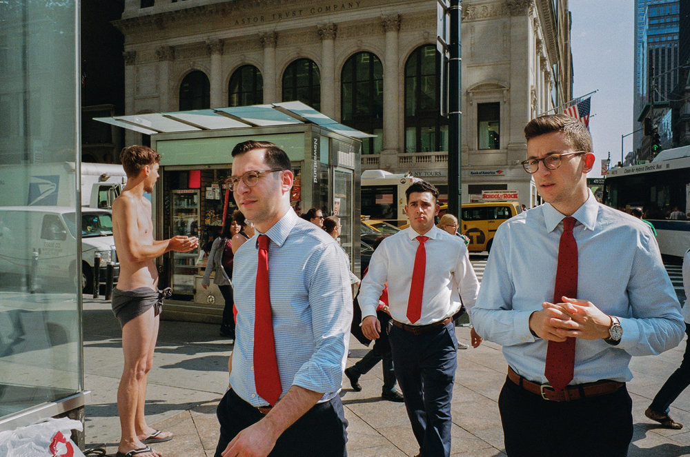 Three similar-looking men in blue shirts and red ties walking toward the camera on a Manhattan street