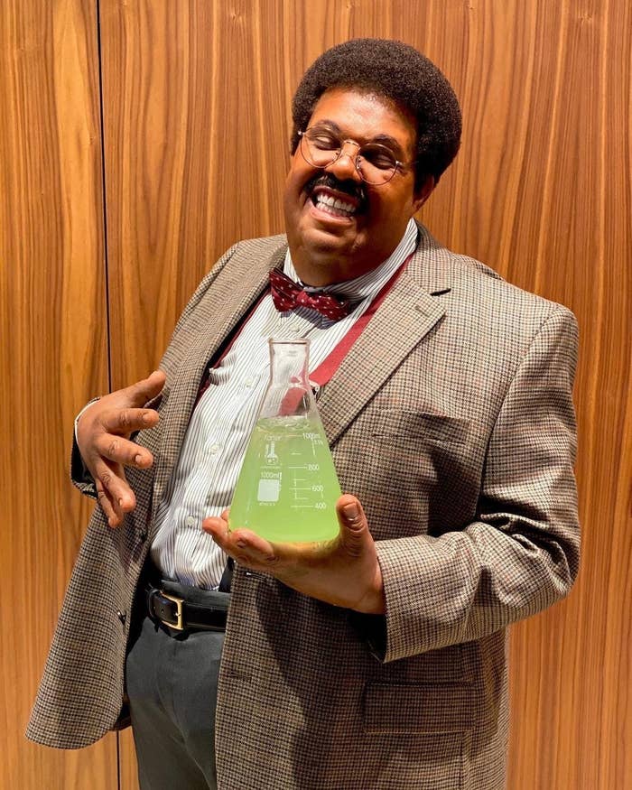 The Weeknd wearing slacks, suspenders, a jacket, and a bowtie while holding an Erlenmeyer flask filled with liquid