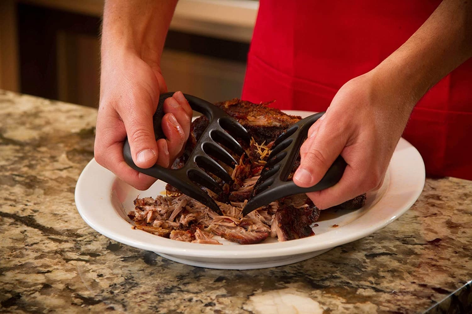 Hands use two black shredding claws to cut up pulled pork on a white plate