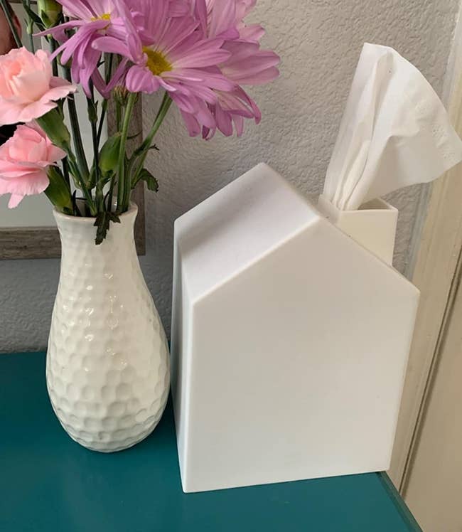 A three-dimensional house-shaped tissue holder sitting next to a vase of flowers 