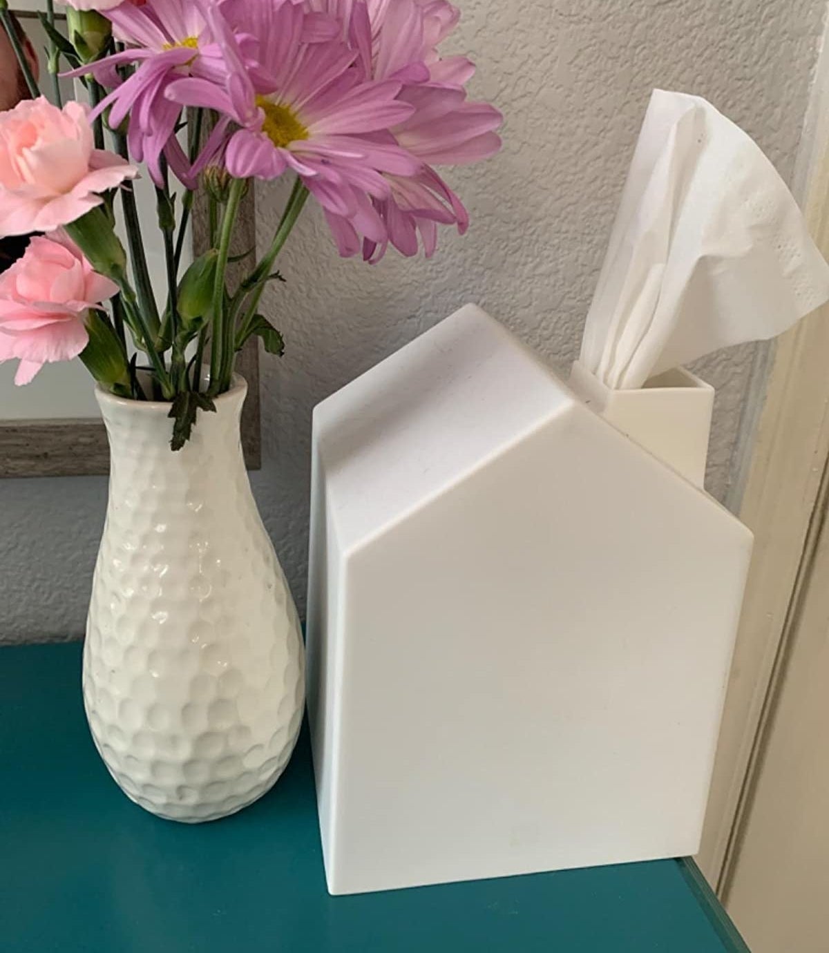 A three-dimensional house-shaped tissue holder sitting next to a vase of flowers 
