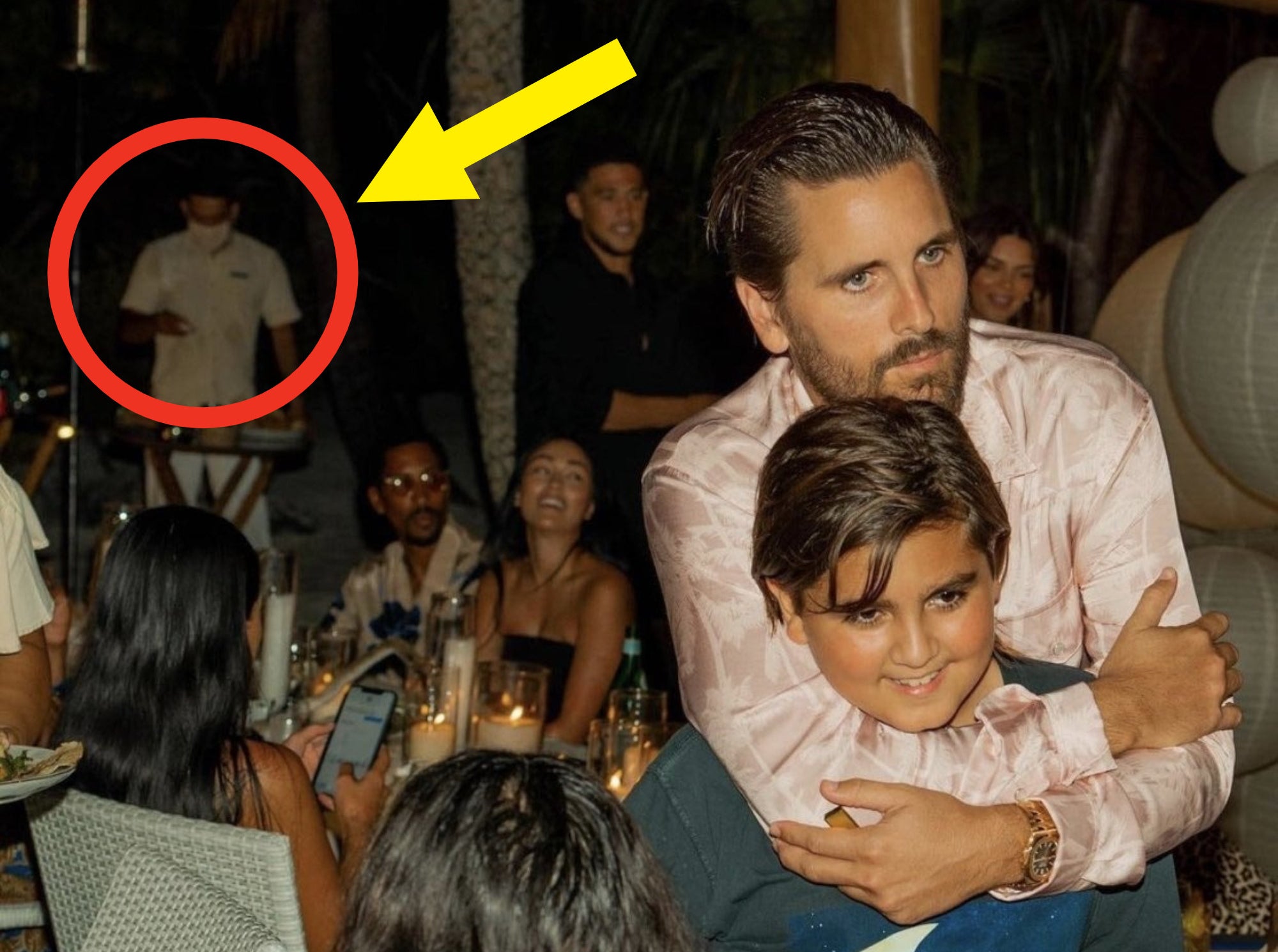 Scott and Mason Disick hugging as a masked waiter works in the background.