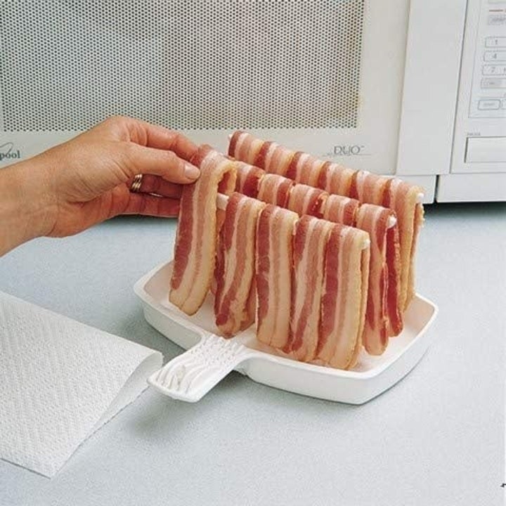 Hand places bacon pieces on white bacon microwave cooker
