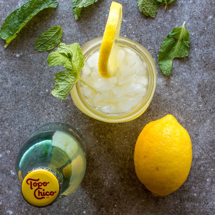 A lemon, mineral water, and mint surrounding a glass of boozy lemonade. 