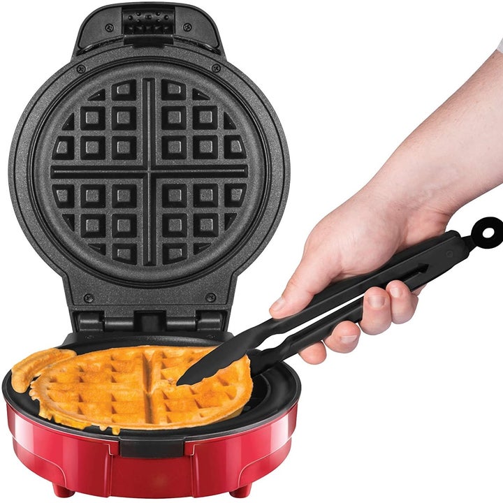 A waffle being taken out of a waffle maker with a pair of tongs