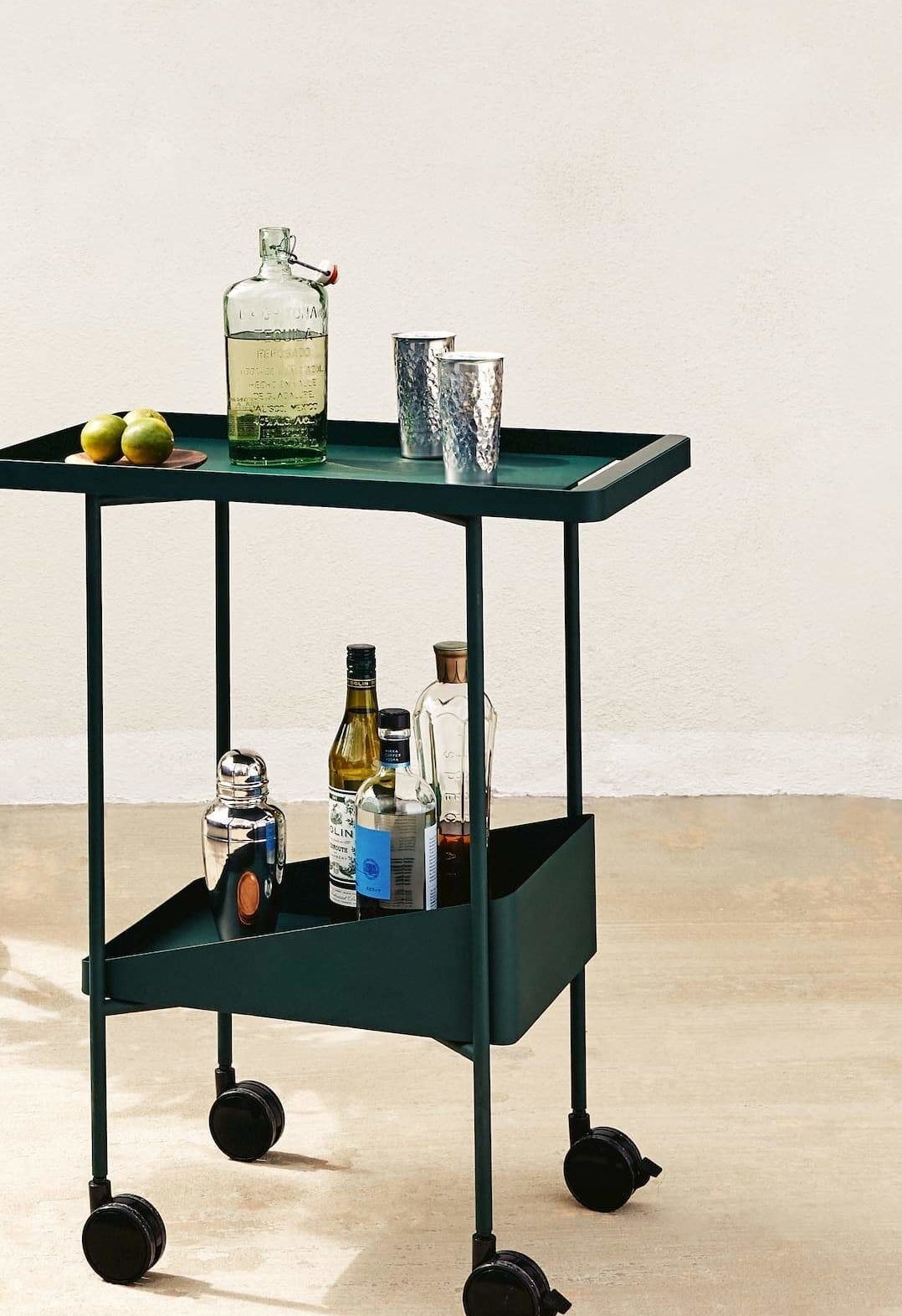 A dark green rolling trolley cart with a serving top and bottom shelf filled with bottles and cocktail supplies
