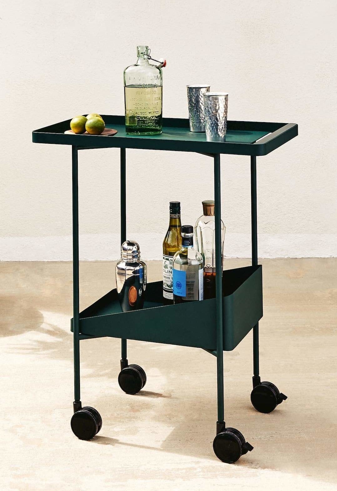 A dark green rolling trolley cart with a serving top and bottom shelf filled with bottles and cocktail supplies