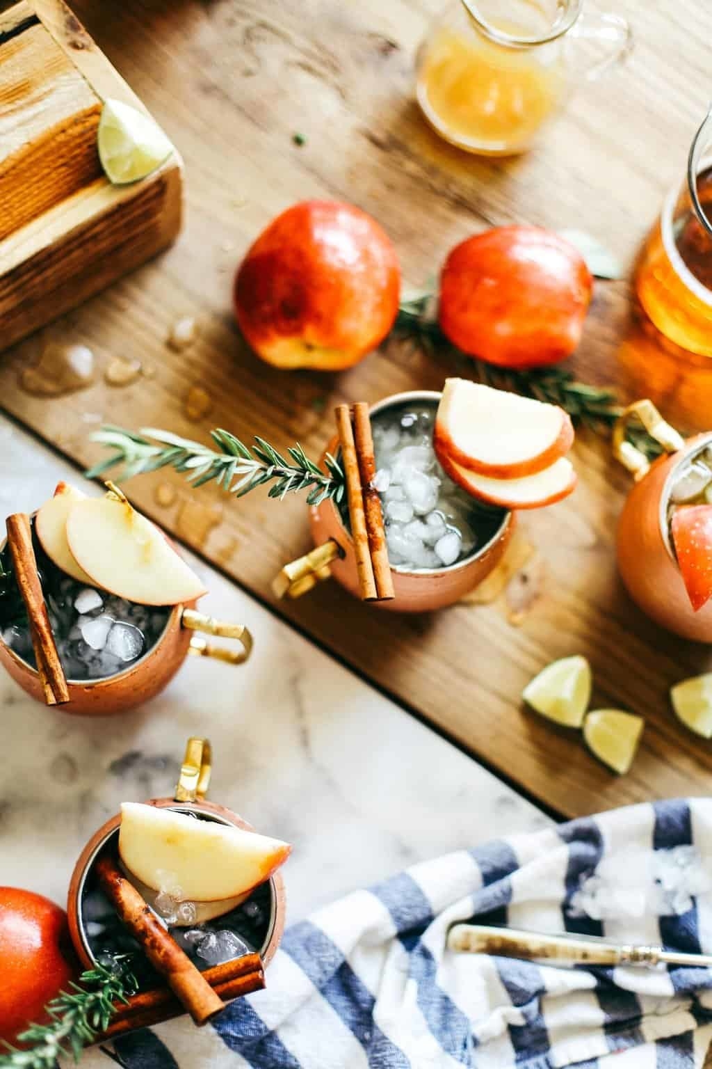 Half a dozen copper mugs filled with apple Moscow mules surrounded by lime wedges, cinnamon sticks, and thyme.