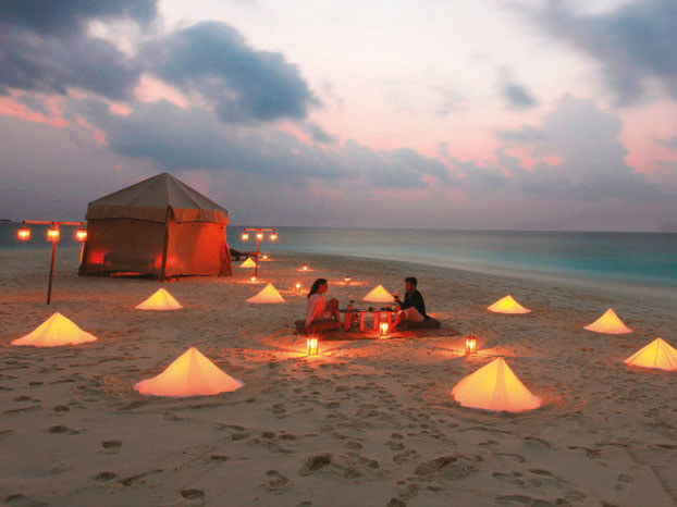 A couple sitting on a deserted beach next to a luxury tent surrounded by lanterns