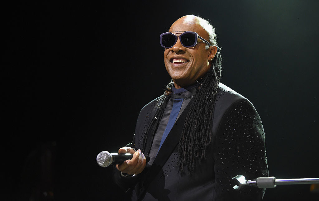 Stevie Wonder performing onstage, holding a microphone and wearing sunglasses