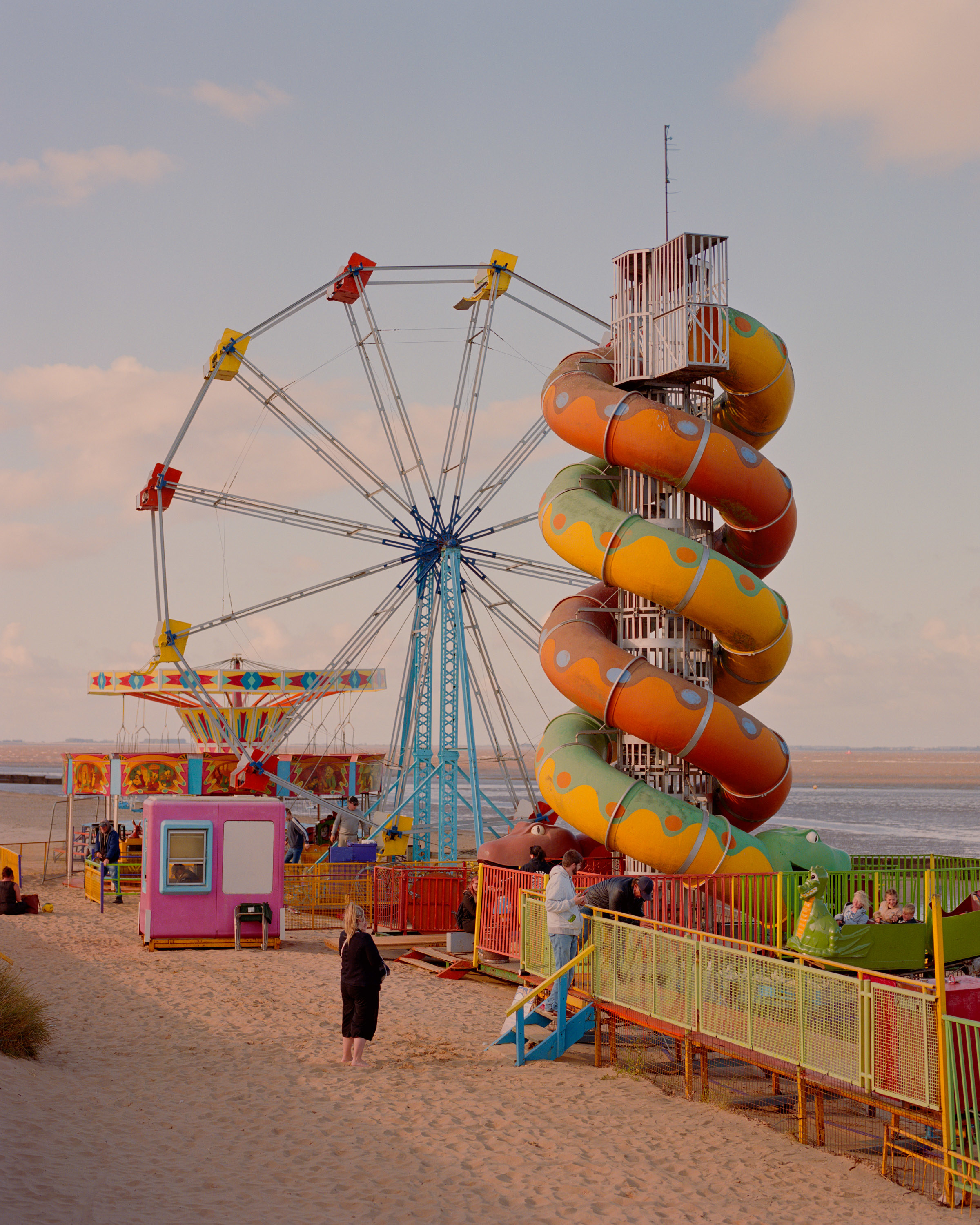 A woman in black and a man in a white sweatshirt stand next to colorful carnival equipment on the beach. 