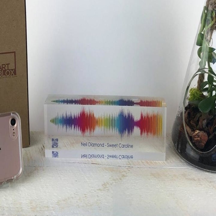 A slim glass block printed with a rainbow soundwave and text that reads "Neil Diamond -  Sweet Caroline" with a small barcode in the bottom-left corner 