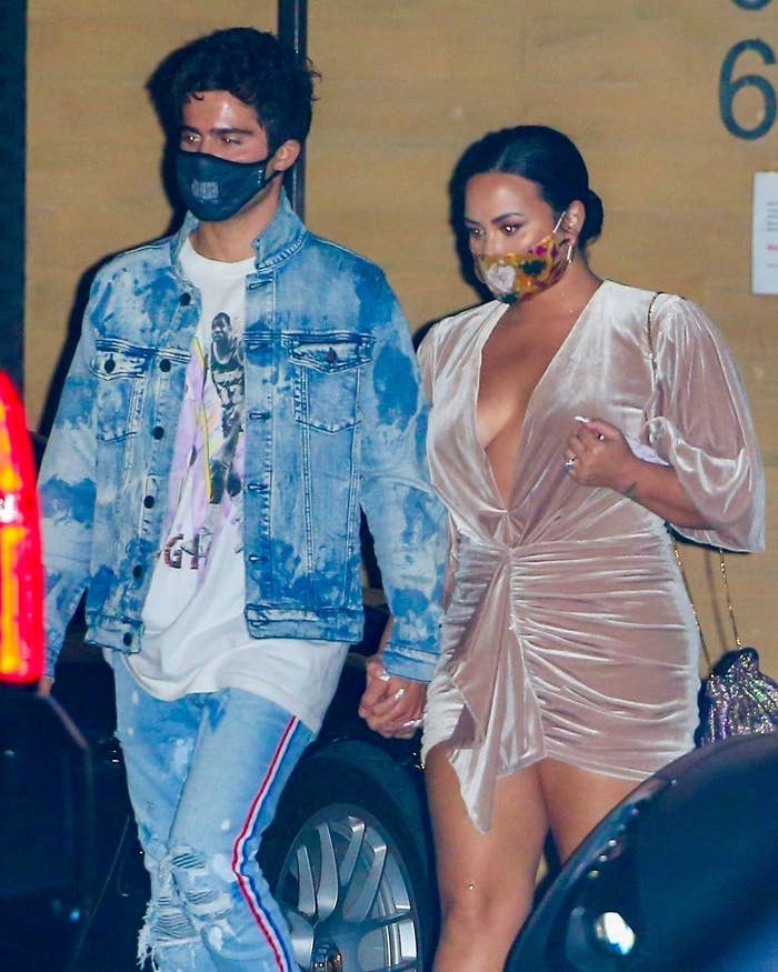 Max, wearing a face mask, T-shirt, jeans, and denim jacket, walking hand-in-hand with Demi, who&#x27;s wearing a short velvet dress