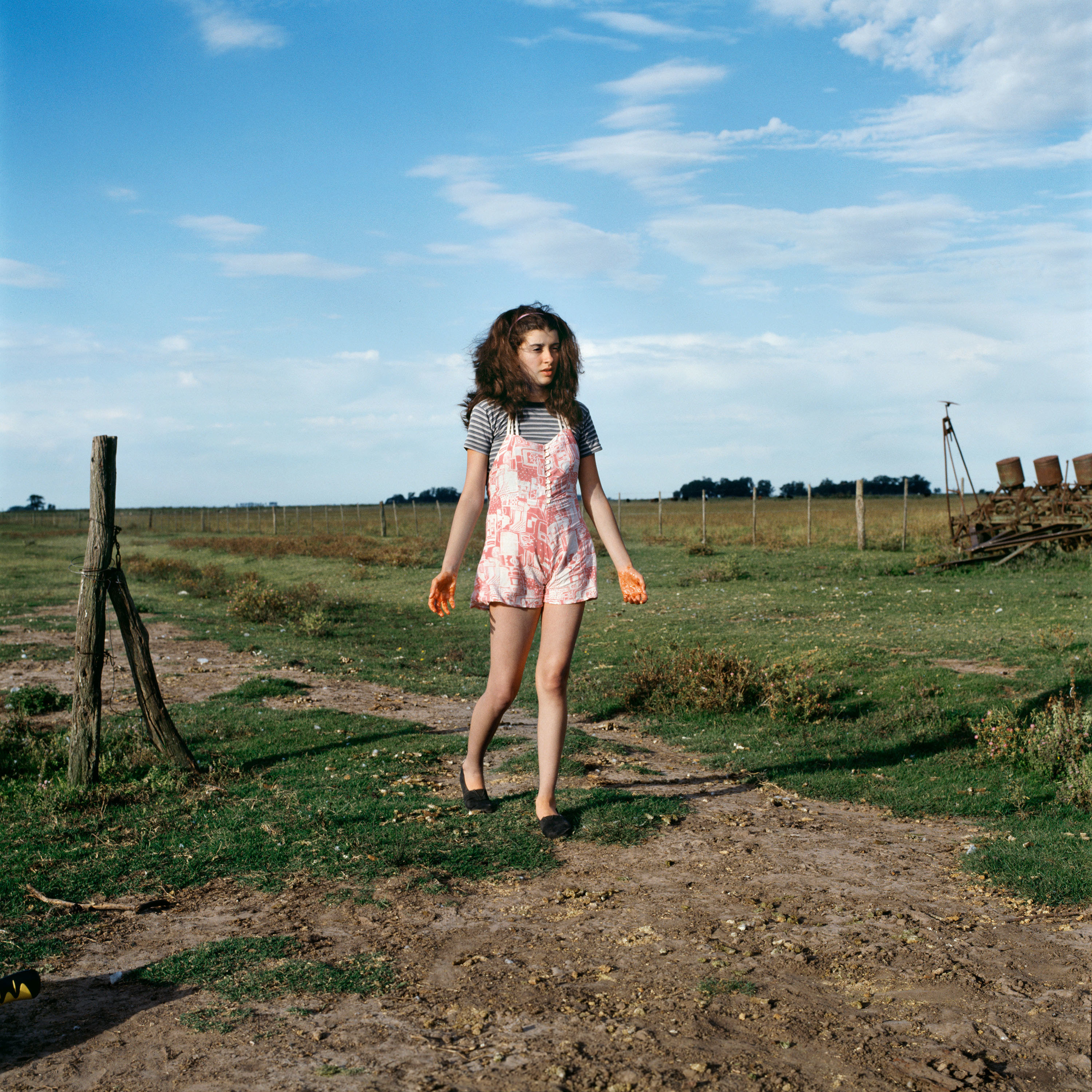 A girl in a striped shirt and pink romper walks through a field under a blue sky