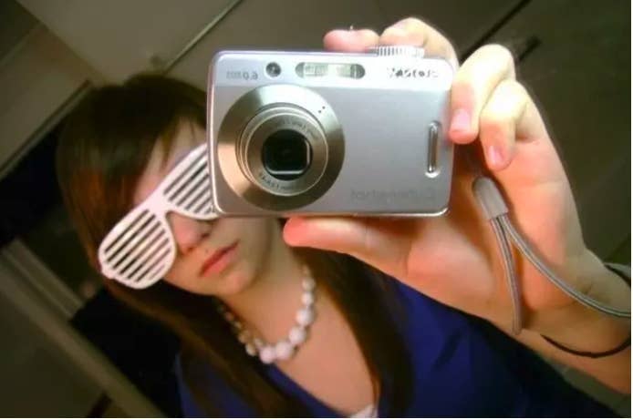 Photo of a girl taking a mirror selfing wearing shutter shades and pointing the camera in front of her face toward the mirror