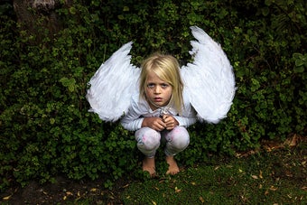 A small child wearing pajamas and angel wings crouching down in from of a hedge