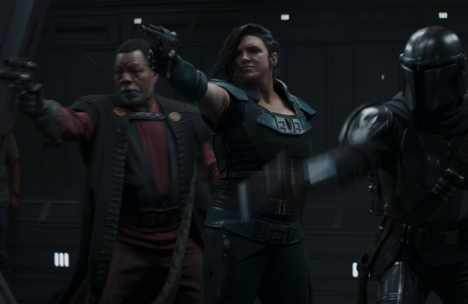 Greef, Cara, and the Mandalorian firing blasters in a corridor with a crew member in jeans and a T-shirt barely visible on the left side of the screen