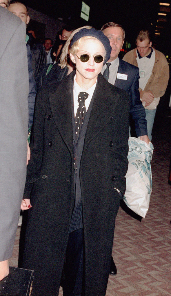 Madonna in a suit jacket with a tie underneath, dark loose pants, and a long peacoat on top