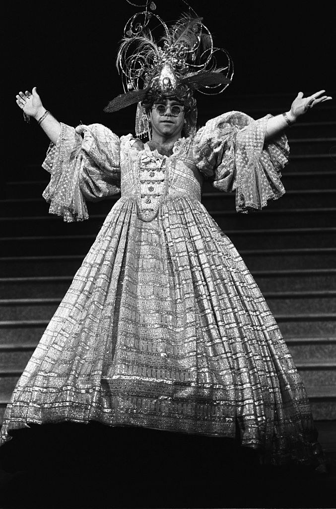 Elton in a large elaborate dress with trailing ruffled sleeves and a poofy skirt and large hat with swirling wire sticking out