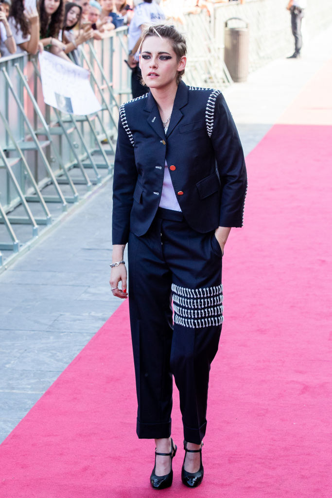 Kristen in loose cropped ankle pants with four layers of hanging chains mid-thigh on one leg and a loose jacket with only the top button fastened with similar chains around the shoulders