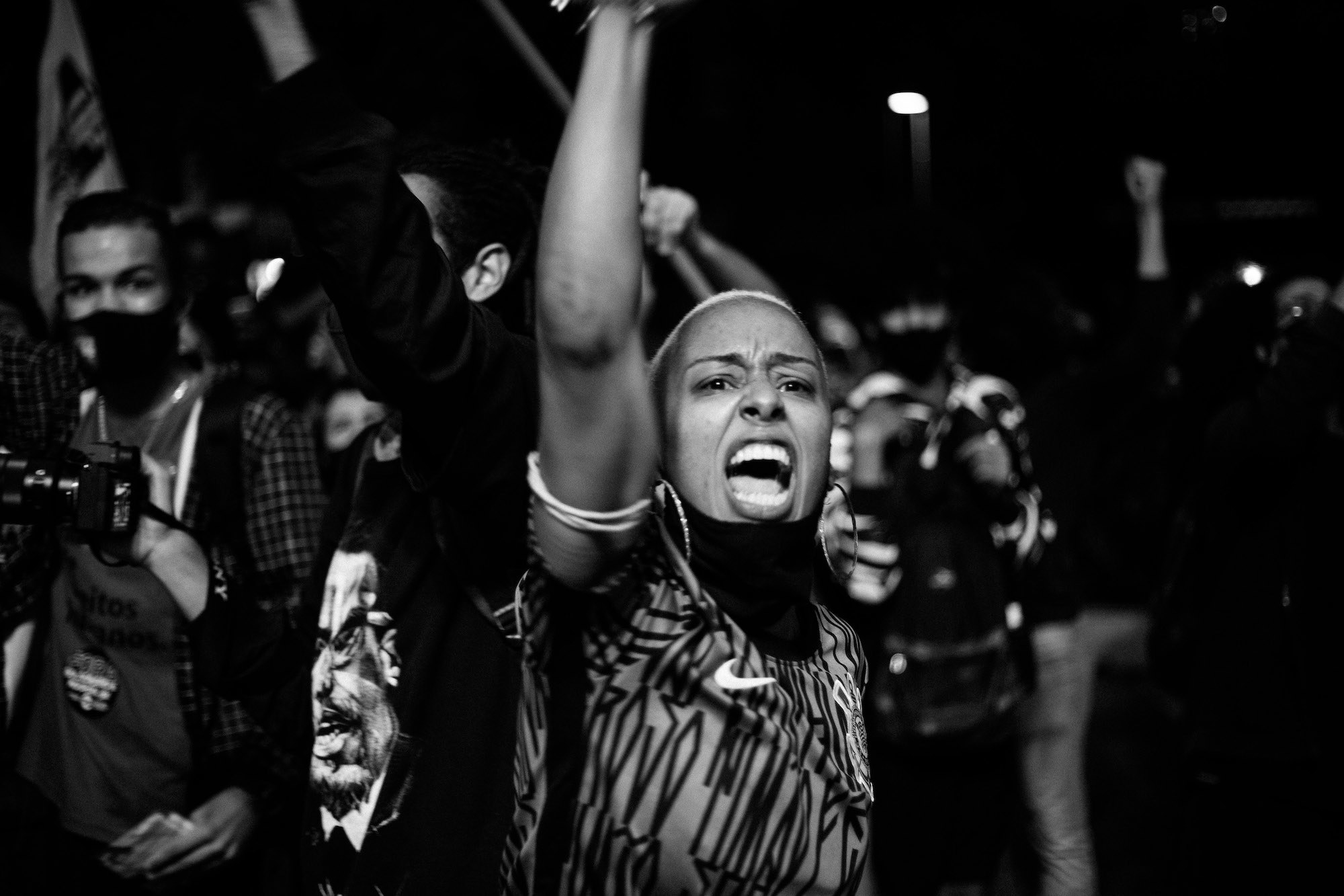 Mulher de raspado vestindo camiseta com estampa de pixo e brincos de argola, com bandana preta no pescoço, gritando com braço erguido, pessoas com braços erguidos passando atrás.