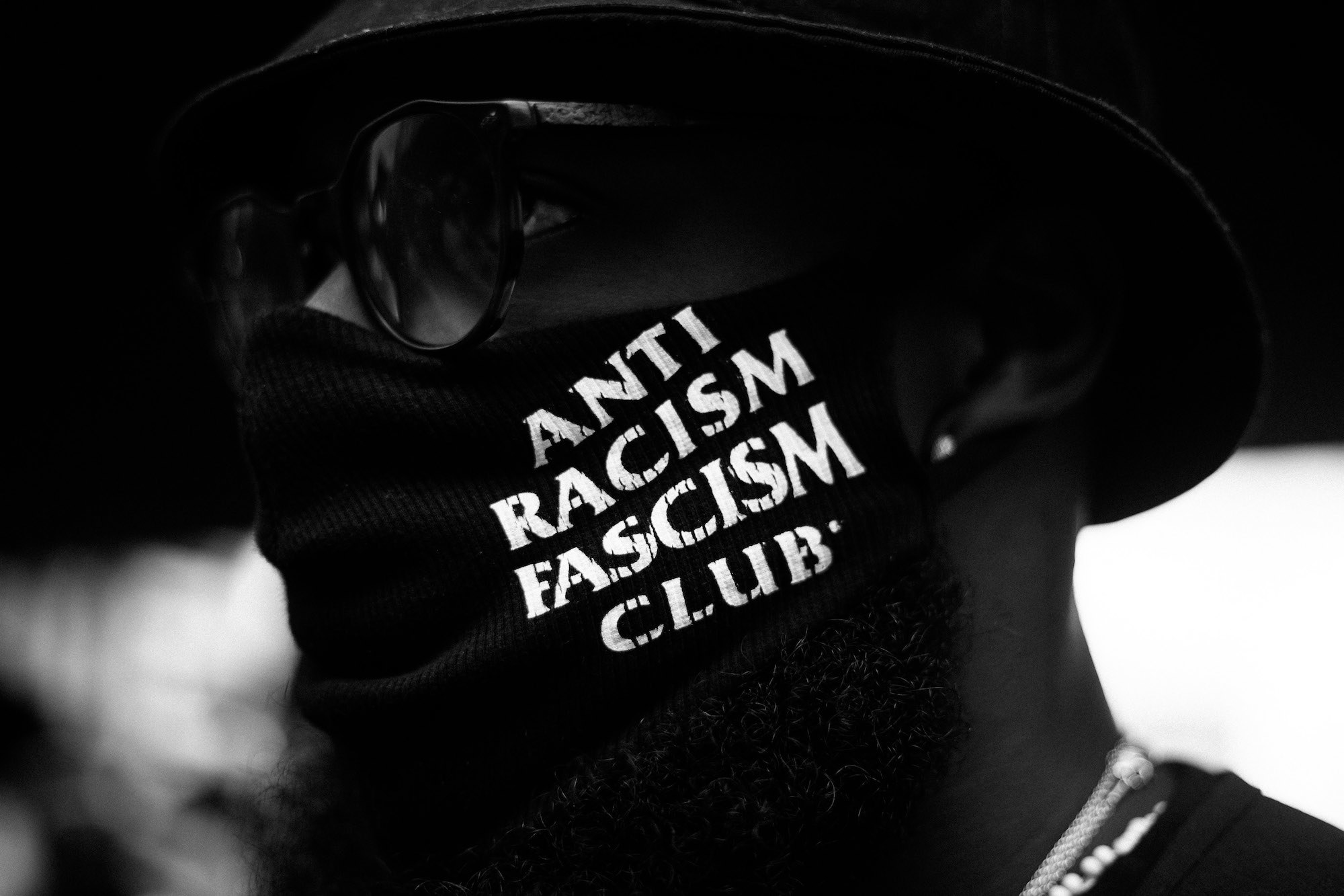 Close de rosto pessoa negra escondido atrás de máscara preta onde se lê em letras brancas ANTI RACISM FASCISM CLUB.