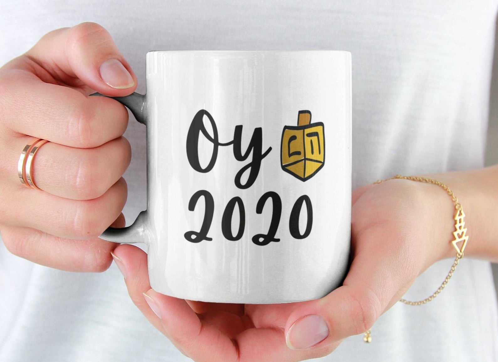 a person holding the white mug that reads &quot;oy 2020&quot; in black text with an illustration of a dreidel