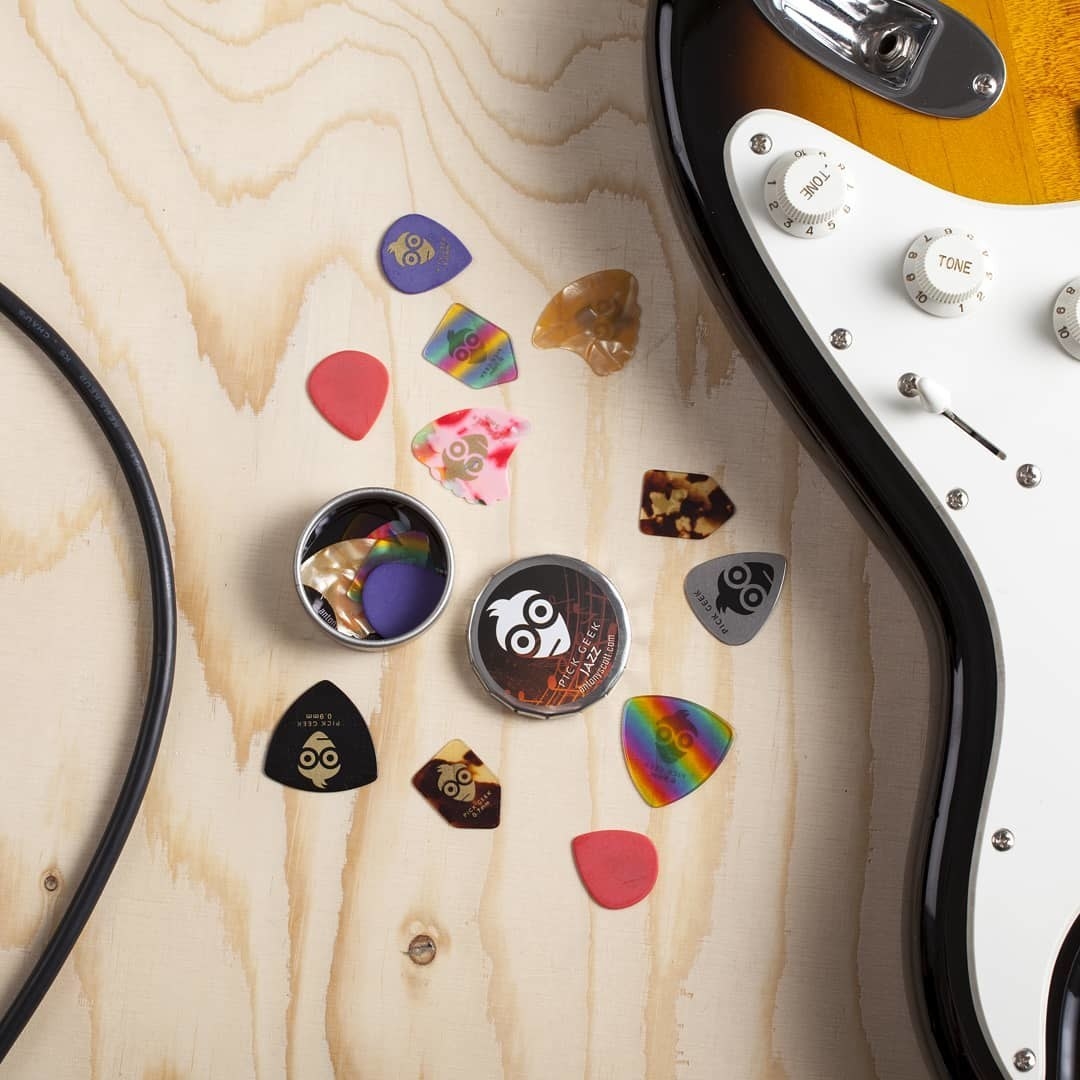 An open tin of various guitar picks arranged on an unfinished wooden surface next to an electric guitar